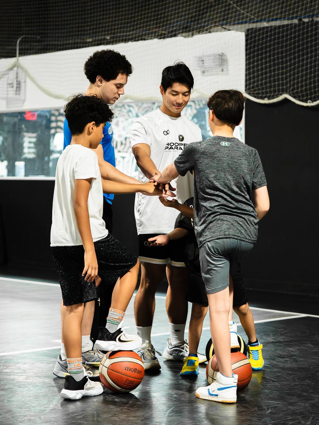 Young players and DETAIL Miami coaches stacking hands in a huddle, basketballs at their feet